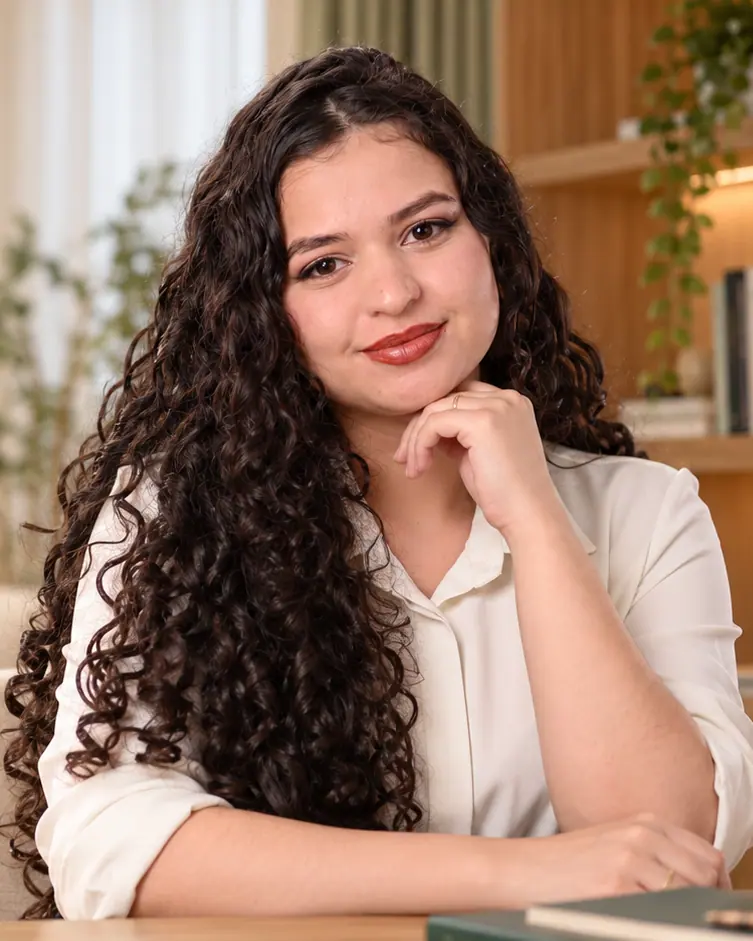 Kimberlly Pablinni, psicóloga, sorrindo com a mão apoiada no queixo, em um ambiente claro com plantas e luz natural